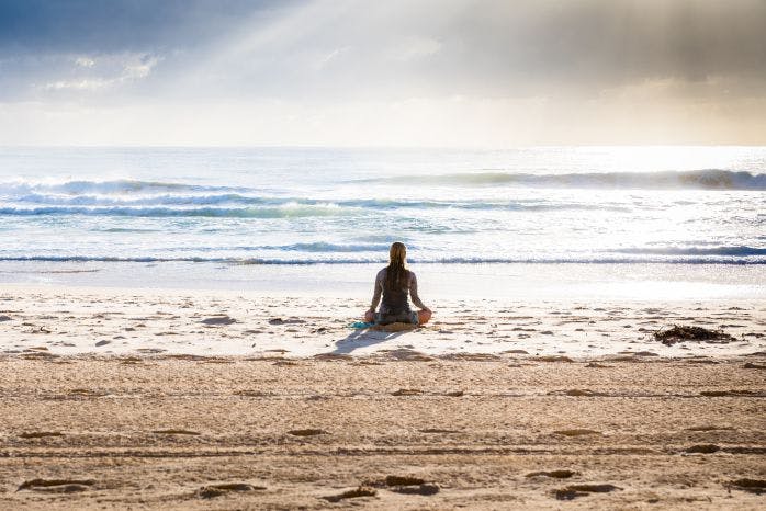 pessoa sentada na áreia na posição de meditação olhando para o mar